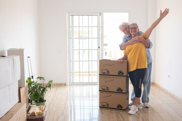 Cheerful senior couple enjoys moving into a new home, surrounded by moving boxes in an empty room. The woman has outstretched arms, symbolizing excitement for their new beginning as retirees