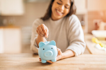 Happy Woman Putting Coin In Piggy Bank At Home Kitchen Table