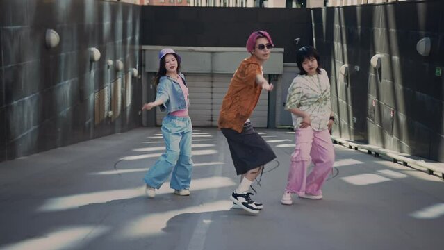 Full shot of young talented k-pop trio consisting of pink haired guy and two pretty girls in vibrant clothes dancing at camera on street