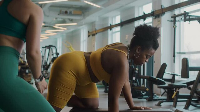 Medium shot of plus size young Black woman in yellow activewear bending over on bench while doing dumbbell row exercise with help of female trainer during gym workout