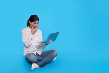 Woman Sitting on Blue Background, Looking at Laptop in Surprise