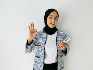 Portrait of smiling Asian Muslim woman OK gesture with the smartphone wearing black hijab and tweed blazer over white background