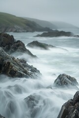 Fototapeta premium Waves crashing against rocks, with a softly blurred background of a rugged coastline and sea. 