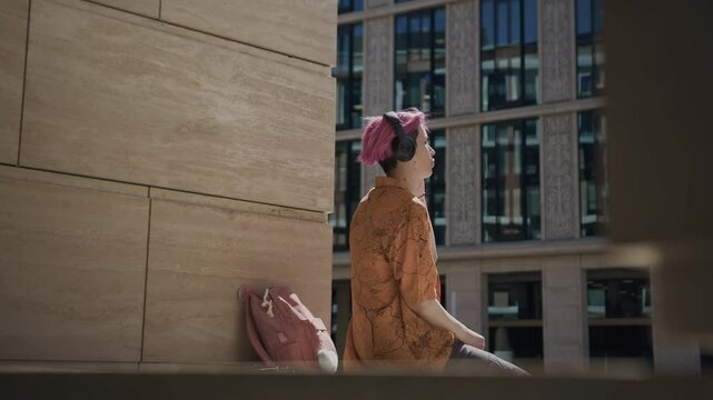 Medium slowmo of Asian guy with pink hair listening to k-pop music in headphones while sitting outdoors under sunlight