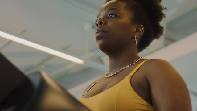 Tilt up shot of determined plus size African American woman in yellow activewear, white socks and sneakers walking on treadmill while working out at gym