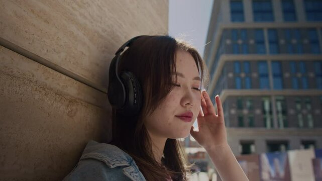 Lens flared slowmo shot of pretty Korean girl in headphones listening to kpop music while sitting outdoors on sunny day