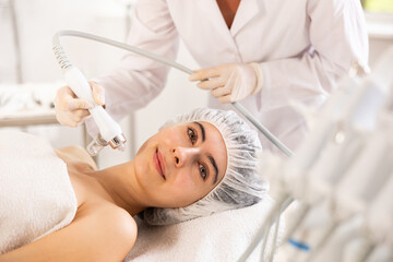 Positive young girl undergoing facial procedure with application of atomized nutrients to enhance skin vitality at aesthetic medicine clinic. Modern technologies in hardware cosmetology