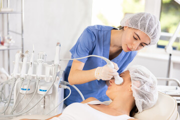 Young female cosmetologist performs hardware facial ultrasound procedure to young female patient