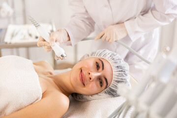 Young woman undergoing skin therapy session in modern cosmetology clinic. Aesthetician applying atomized nutrients with airbrush onto client face to enhance and revitalize complexion