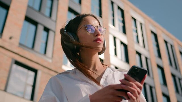 Happy carefree hispanic woman listening music in headphones on phone using mobile audio application in city outdoors sunshine relaxed girl female businesswoman student relaxing enjoying mp3 on street