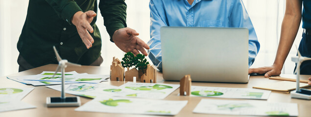Cropped image of skilled businessman presented eco-friendly house project to general manager at meeting table with wooden block, windmill model and environmental document scatter around. Delineation.