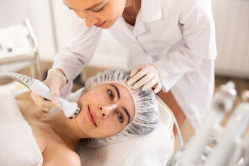 Young female cosmetologist performs hardware facial ultrasound procedure to young female patient