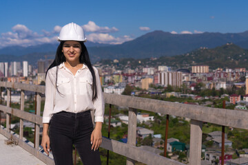 Young brunette resident architect in a hard hat on a construction site against the background of the city and mountains on a sunny day. Successful inspector.