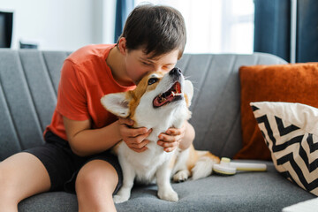 Friendly teenager with down syndrome embracing and kissing his corgi dog on comfortable sofa