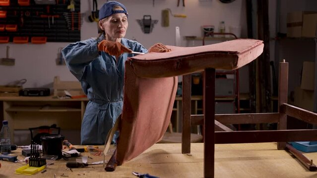 In a home workshop, a woman uses a chisel to remove the backrest from an old wooden chair.