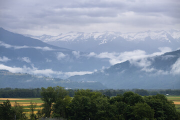 Obraz premium A scenic of mountain with layers covered with clouds, fog and snow with cloudy sky after rain in Savoie region of France . A greenery nature surrounded with French agriculture. 