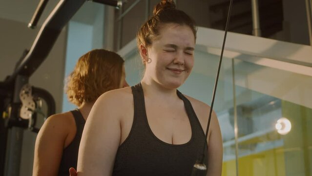 Medium shot of hard-working young Caucasian plus size woman doing triceps pushdowns while training with female coach at gym