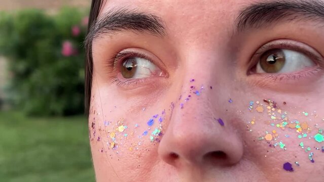 Horizontal video close up. Brown eyes of young adult white woman look away. Female face with multicolored purple, gold, blue, green sparkling glitter on her cheeks. Rest, relax, march 8, mental health