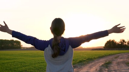Girl standing on rural dirt road in field with green grass. Raises hands up saying goodbye to sunset. Children stretches arms out to sides meeting dawn morning. Does exercises and charged heat energy
