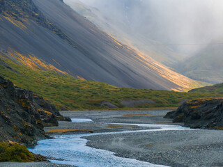 A winding river flows through a rocky valley in Iceland, bordered by steep mountains. The sun...