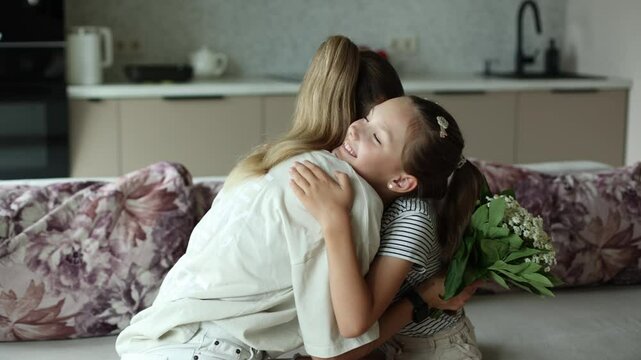 Child daughter is congratulating mom and giving her flowers. Mum and girl smiling and hugging, mother kisses a daughter