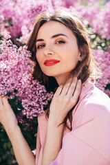 Young beautiful smiling woman. Sexy carefree model posing in the street. Positive model near blooming flower lilac bush beaming. Summertime, pink colours. Closeup beauty portrait, red lips