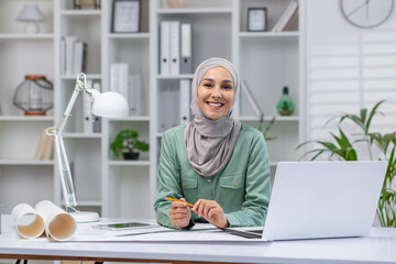 Smiling female architect wearing hijab working at modern office desk with laptop, blueprints, and tablet. Woman in office workspace. Concept of female empowerment, business, and architecture.