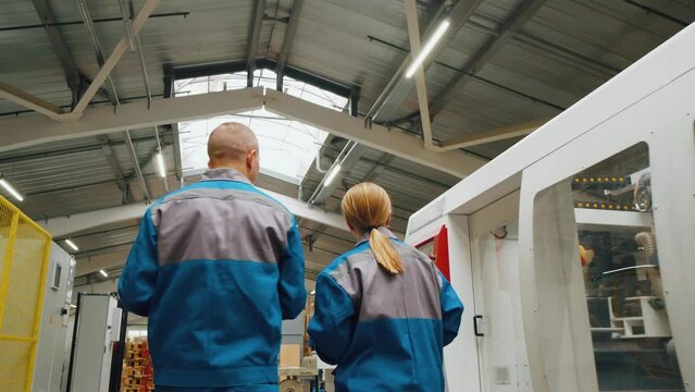 Two engineers technicians workers walk through production workshop of a modern factory. Workers actively discuss work process, machines, equipment. A man and a woman are employees of a huge factory.