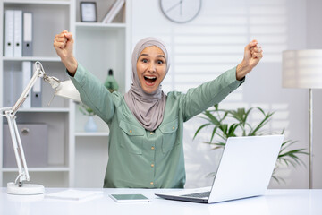 Joyful woman in hijab cheering at desk with laptop, celebrating professional achievement in bright...