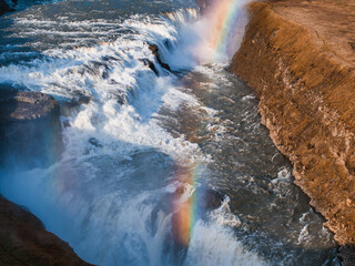A stunning waterfall in Iceland cascades over a dark brown rocky cliff, creating a light blue spray. A vibrant rainbow arcs above, adding to the breathtaking scene.