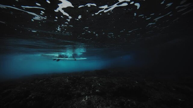 Underwater angle sideview of surfer duck diving and pushing down on board as wave crashes