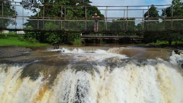 Revealing Drone Shot of Woman Standing on Footbridge Above Scenic Waterfall on Sunny Day, Mena Creek Falls, Australia