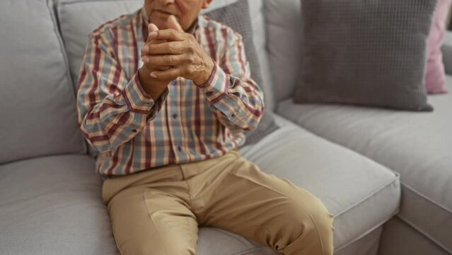 An elderly hispanic man with grey hair in a plaid shirt sits on a grey sofa in a living room, holding his hands in a contemplative manner.