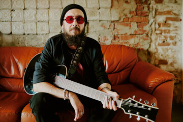 Mature Caucasian rock musician wearing stylish red sunglasses sitting relaxed on leather couch...