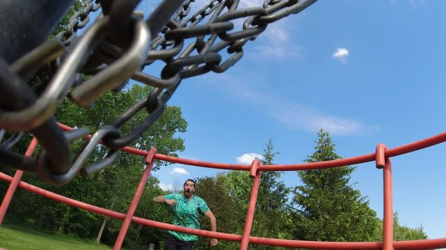 A unique perspective from inside the disc golf basket captures a man celebrating a successful shot, set against a clear blue sky.