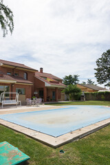 House with garden, tables, chairs and a pool covered with a blue tarp on a sunny day.  End or beginning of season, autumn or spring