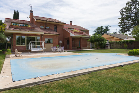 Nice house with green garden with trees and plants surrounding a pool covered with blue tarp protecting the water from dirt on a sunny day. End or beginning of season, fall or spring