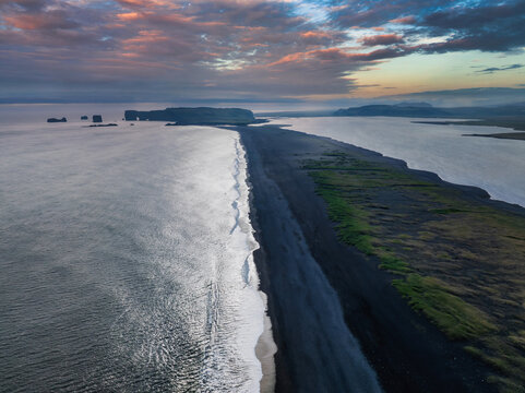 Stunning aerial shot of Iceland's black sand beach with white waves, green vegetation, and a rocky peninsula under a pink and blue sky at sunset.
