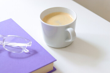 A purple mock up book with transparent glasses and a white cup of coffee on white table. Free copy space for text