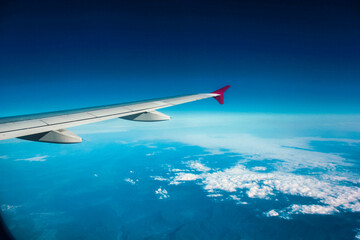 Mid-flight wing view from airplane cabin, highlighting the red winglet. Underneath, vista of land, water, and cloud-dotted sky portrays serenity in air travel.