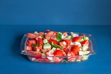 A healthy mix of fresh vegetables seasoned with black pepper and olive oil presented in a glass dish on a blue background with ample copy space, making the vibrant veggies stand out. Healthy eating.