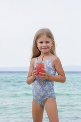 Adorable little girl in blue swimwear and black sunglasses having refreshment drink on the sand...