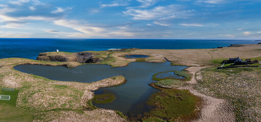 Stunning aerial view of Iceland's coastline featuring deep blue lakes and ponds, surrounded by green grass and brown rock under a bright blue sky with white clouds.