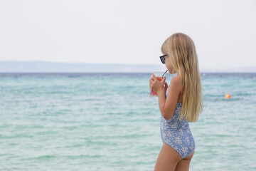 Adorable little girl in blue swimwear and black sunglasses having refreshment drink on the sand...