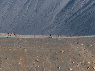 A small figure walks along a smooth path at the base of a dark gray, textured crater in Iceland. The crater's rough surface dominates the landscape.