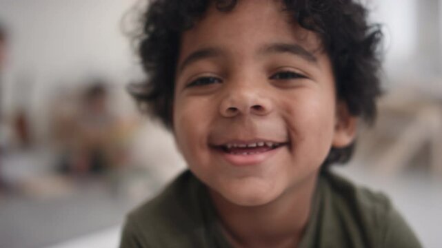 Close-up portrait of happy African American boy with curly hair smiling for camera while spending time in kindergarten, blurry nursery interior in background
