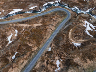 Aerial perspective of a paved road winding through a rocky, barren landscape in Iceland. Small streams and patches of green vegetation add contrast to the scene.