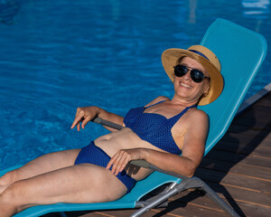 An old woman in a hat and sunglasses is sunbathing on a sun lounger by the pool. Vacation in retirement. 