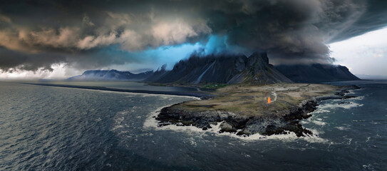 A dramatic dark sky with swirling clouds hangs over a Vestrahorn mountaine on Stokksnes cape in Iceland. Deep blue waves crash against the shore, with a small orange structure visible in the distance.