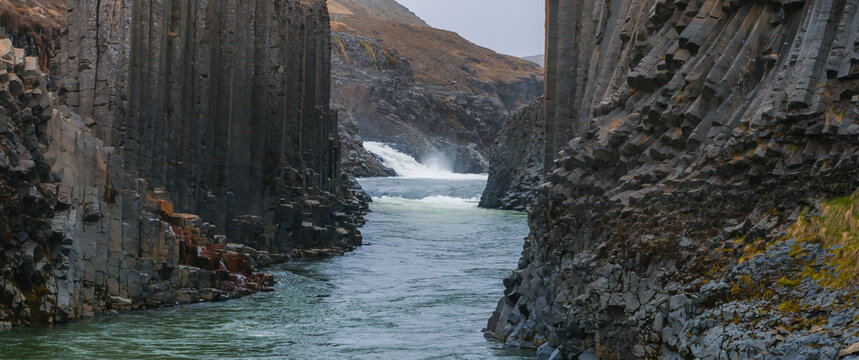 Dramatic aerial shot of a whitewater river cutting through a narrow canyon with steep, dark rock formations in Iceland. The contrast creates a stunning visual.